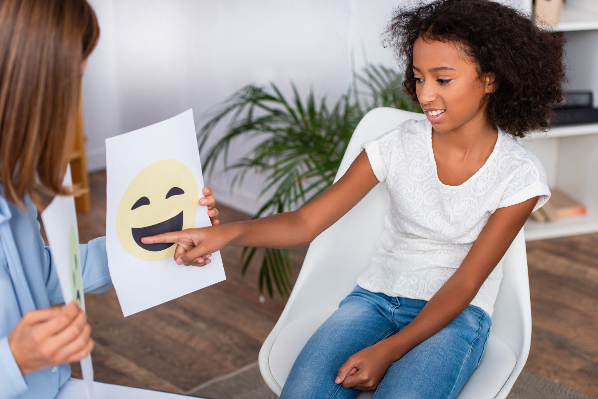 Smiling african american girl pointing with finger at happy expression on paper near psychologist on blurred foreground