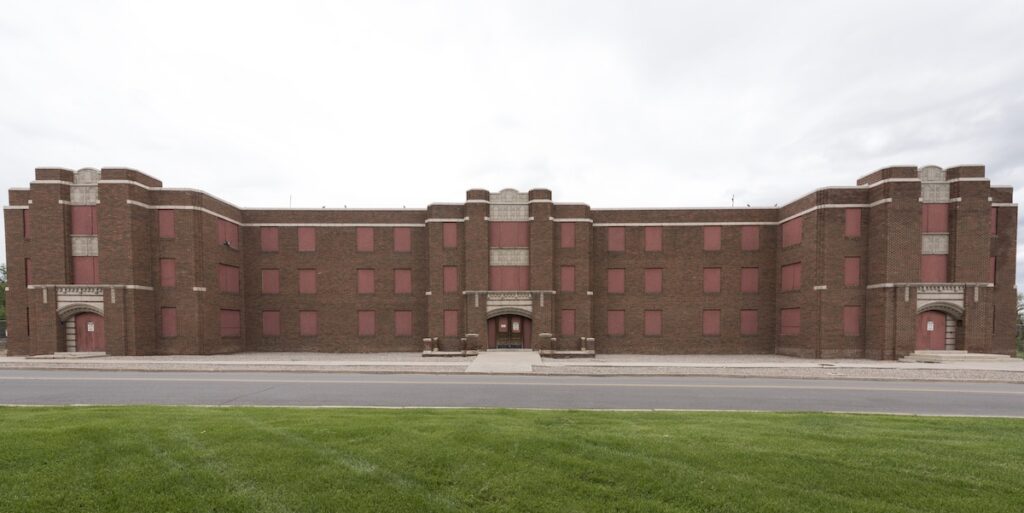 Front view of a large, symmetrical, abandoned four-story brick institutional building with boarded red window openings and three recessed entrances. The structure spans the width of the image, facing an empty roadway, with a green lawn in the foreground and an overcast gray sky overhead. Architectural details include decorative brickwork, arched entryways, and a central entrance flanked by projecting corner sections.