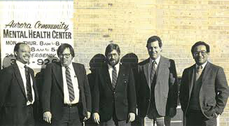 Historic black-and-white group photograph of five people standing in front of a brick building. A painted sign on the wall reads “Aurora Community Mental Health Center” with additional smaller text and a phone number partially visible. The individuals are dressed in business attire, including suits, ties, and blazers, and are arranged in a row facing the camera. The image has a vintage appearance with visible grain and slight fading.