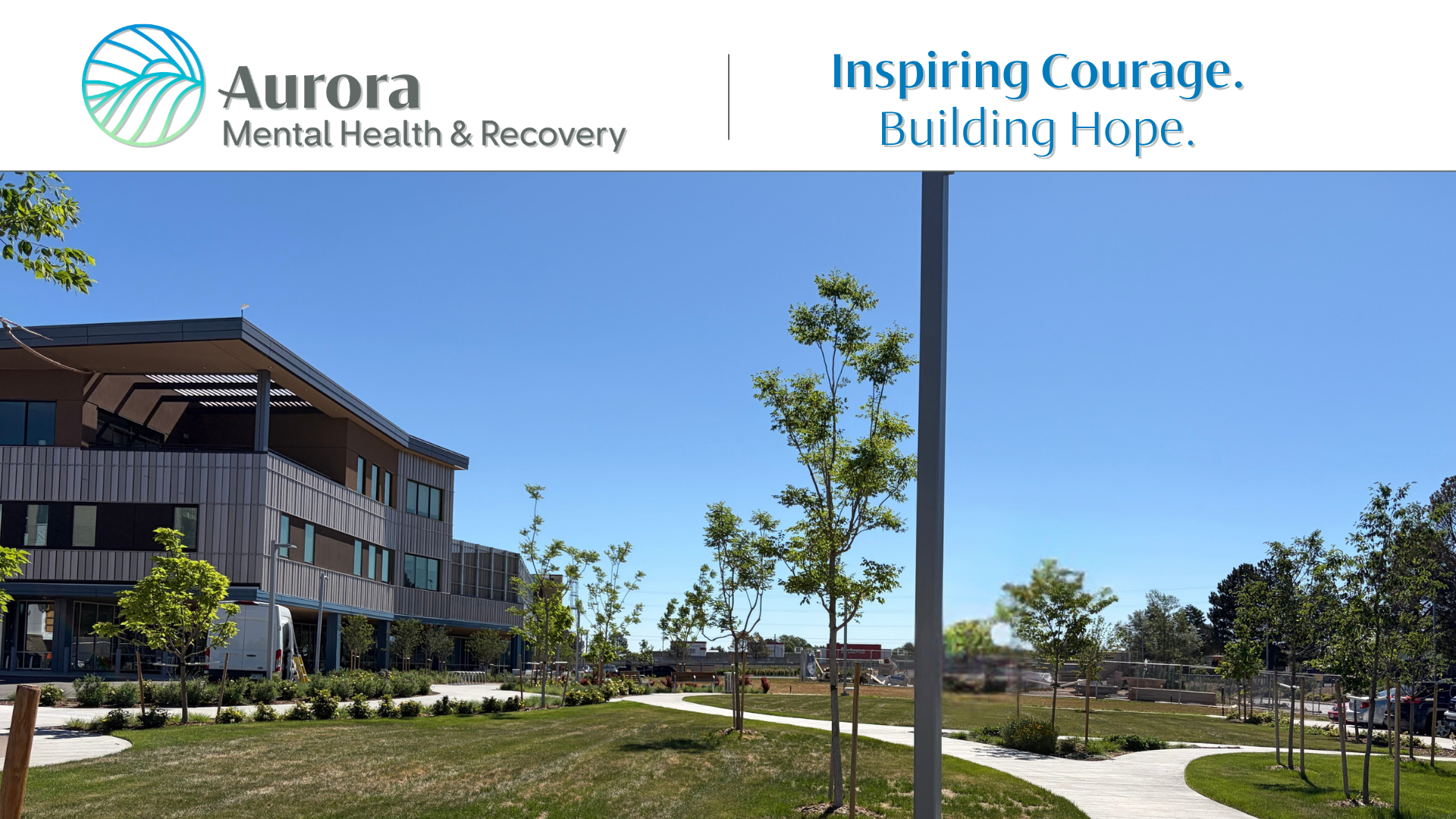 Banner image for Aurora Mental Health & Recovery. The top section displays the organization’s logo and name on the left and the tagline “Inspiring Courage. Building Hope.” on the right. Below, a wide photo shows a modern healthcare campus with a three-story building, landscaped lawn, young trees, and winding sidewalks under a clear blue sky.