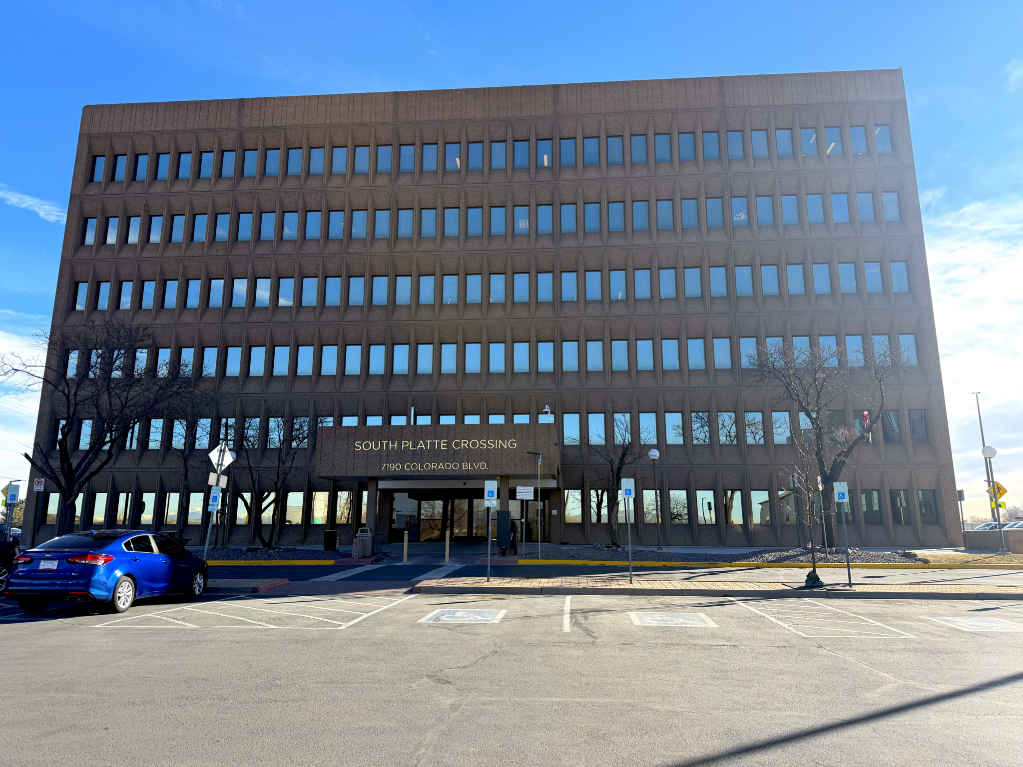 Exterior view of the South Platte Crossing office building at 7190 Colorado Blvd. A six-story brown office building with rows of blue-tinted windows stands behind a parking lot with accessible parking spaces. A blue car is parked in front, with leafless trees framing the entrance under a bright blue sky.